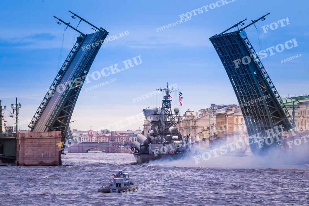 Фотография демонстрирует момент, когда военный корабль проходит под разведённым мостом на реке Нева в Санкт-Петербурге. Это изображение запечатлело динамичную сцену с участием военной техники на фоне исторической архитектуры города. Фотография подходит для использования в публикациях о военно-морской тематике, истории Санкт-Петербурга или туристических гидах.